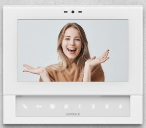 Schermo del videocitofono Klass di Videx con un'immagine di una donna sorridente. Il dispositivo è caratterizzato da un design elegante con finitura in vetro, spessore ultrapiatto, e un'interfaccia touch con icone luminose per le funzioni principali